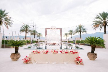 Çiçek buketleriyle dolu uzun festival masası, kenarında yeşil palmiye ağaçları olan bir havuzun önündeki hasır heykelin yanında duruyor. Yüksek kalite fotoğraf
