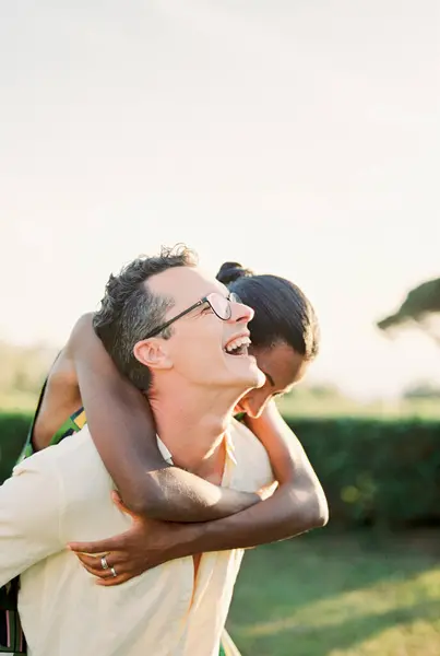 Laughing man carries woman on his back across green lawn. High quality photo