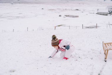 Küçük kız, karlı bir açıklıkta tahta bir kızağın yanında çömelirken kartopu yapıyor. Yüksek kalite fotoğraf