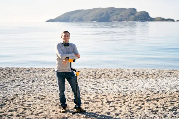 Gülümseyen adam kıyıda metal dedektörüne yaslanmış duruyor. Yüksek kalite fotoğraf