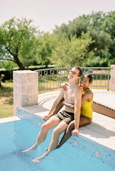 Smiling woman hugs man from behind while sitting with him on the edge of a swimming pool. High quality photo