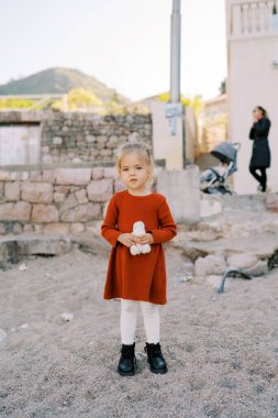 Ellerinde yumuşak bir oyuncak olan küçük bir kız sahildeki taş bir çitin yanında duruyor. Yüksek kalite fotoğraf