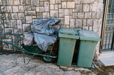 Cart with bags of garbage stands near plastic garbage containers near a stone fence. High quality photo clipart