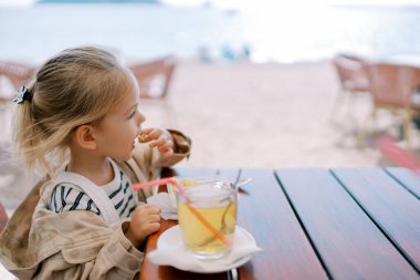 Küçük kız plajda bir bardak meyve suyunun yanında pipetle otururken yemek yiyor. Yan görüş. Yüksek kalite fotoğraf