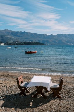 Sandalyeli ahşap masa, deniz kenarındaki kumlu bir sahilde demirli kırmızı bir tekneyle duruyor. Yüksek kalite fotoğraf