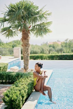 Young woman sits on the edge of a swimming pool, hugging her knee and looking away. High quality photo clipart