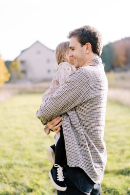 Gülümseyen baba yeşil bir çayırda kollarında küçük bir kızla yürüyor. Yan görüş. Yüksek kalite fotoğraf