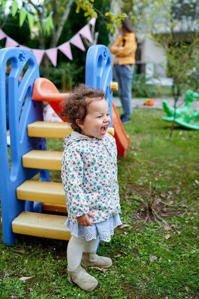 Little laughing girl stands near a colorful slide in the garden. High quality photo — Stock Image Little Laughing Girl Stands Colorful Slide Garden High Quality Photo Stock Image