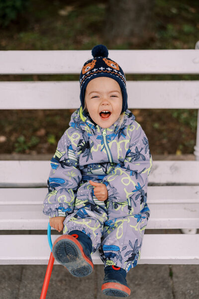 Little laughing girl with a toy in her hand sits on a white wooden bench. High quality photo — Stock Image Little Laughing Girl Toy Her Hand Sits White Wooden Bench Stock Image