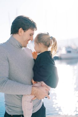 Gülümseyen baba, deniz kıyısında kollarında oturan küçük bir kıza bakıyor. Yüksek kalite fotoğraf