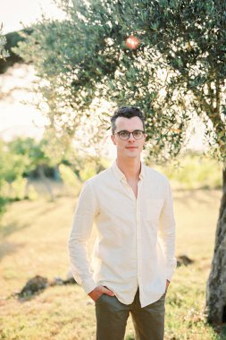 Elleri pantolonunun cebinde genç bir adam bahçedeki ağacın yanında duruyor. Yüksek kalite fotoğraf