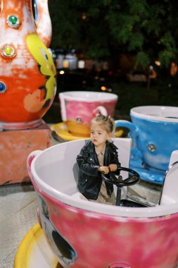 Küçük kız bardak şeklinde bir atlıkarıncanın gidonunu tutarak oturur ve başını çevirir. Yüksek kalite fotoğraf