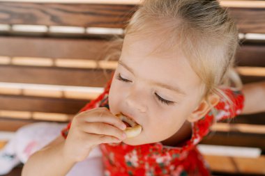 Küçük kız ahşap bir bankta otururken simit kemiriyor. Yüksek kalite fotoğraf