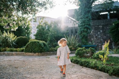 Küçük kız güneşli bir parkta yol boyunca yürür ve yeşil çalılara bakar. Yüksek kalite fotoğraf