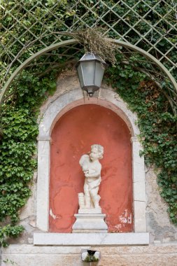 Sculpture of a little boy with an amphora on his shoulder in an arch in a green garden. High quality photo clipart