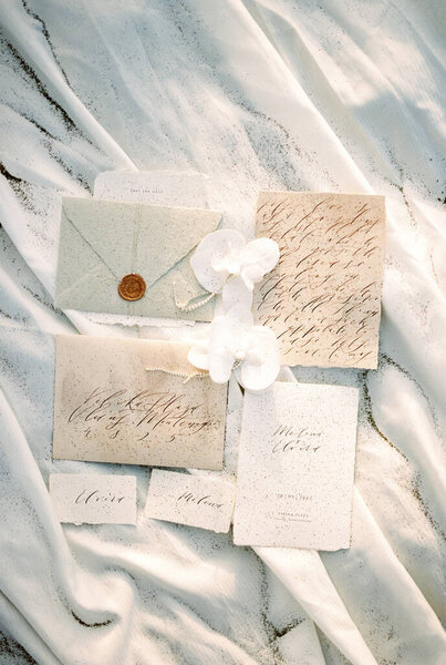 Свадебные приглашения и именные карточки лежат на белой ткани, посыпанной песком. Высокое качество фото