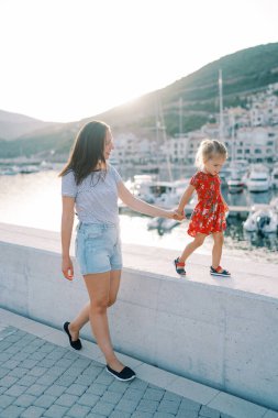 Gülümseyen anne deniz kıyısında yürüyor, küçük kızını kaldırım kenarına götürüyor. Yüksek kalite fotoğraf