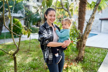 Kollarında küçük bir kızla gülümseyen anne çiçek açan bir bahar bahçesinde duruyor. Yüksek kalite fotoğraf