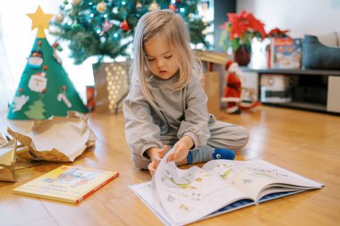 Noel ağacının yanında otururken paketlenmemiş bir kitabı karıştıran küçük bir kız. Yüksek kalite fotoğraf