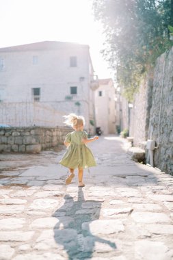 Uçan saçlı küçük bir kız kaldırımlı bir cadde boyunca taş bir çitin önünden geçiyor. Arkadan bak. Yüksek kalite fotoğraf