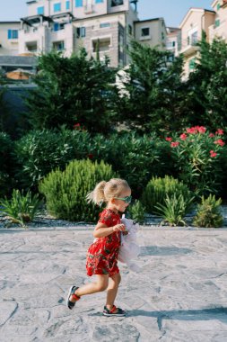 Küçük kız, yerleşim yerlerinin yakınındaki çiçek açan bir bahçede asfalt bir yol boyunca koşuyor. Yüksek kalite fotoğraf