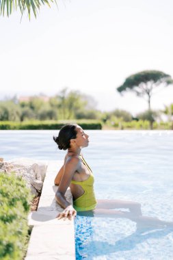 Young woman sunbathes while sitting in a swimming pool and leaning her elbows on the side. High quality photo clipart