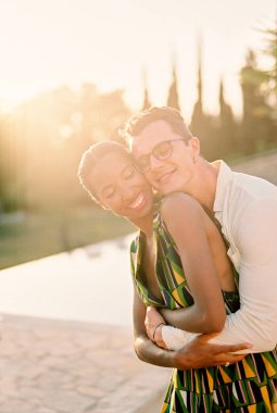 Gülümseyen adam kadına arkadan sarılıyor, güneşli bir parkta dikilirken yanaklarına dokunuyor. Yüksek kalite fotoğraf