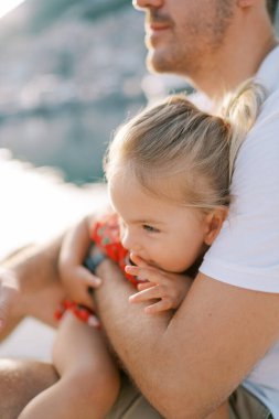 Küçük gülümseyen kız babasının kucağına oturur, eline yaslanır ve mesafeye bakar. Kırpılmış. Yüksek kalite fotoğraf