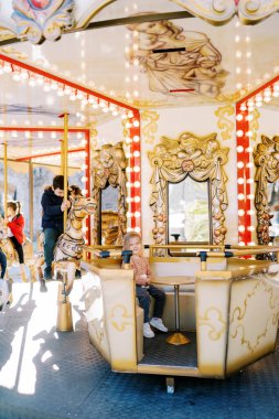 Küçük gülümseyen kız oyuncak atların üzerinde oturan çocukların olduğu bir fuarın atlıkarıncasında bir masada oturuyor. Yüksek kalite fotoğraf