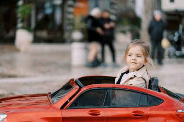 Küçük kız meydanda kırmızı bir oyuncak arabayla dolaşıp yan tarafa bakıyor. Yüksek kalite fotoğraf