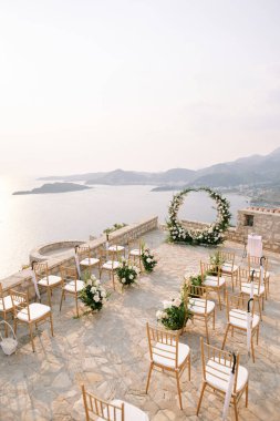 Çiçek buketleri ve katlanmış şemsiyelerle süslenmiş sıra sıra sandalyeler denizin üzerindeki bir gözlem güvertesinde yuvarlak bir düğün kemerinin önünde duruyor. Yüksek kalite fotoğraf
