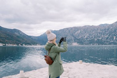 Sırt çantalı bir kız rıhtımda duruyor ve akıllı telefonuyla dağların ve denizin fotoğraflarını çekiyor. Yan görüş. Yüksek kalite fotoğraf