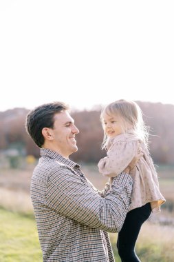 Gülümseyen baba çimlerin üzerinde dururken kollarını açmış gülen küçük kızı tutuyor. Yüksek kalite fotoğraf
