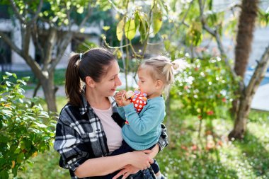 Gülümseyen anne bahçede dururken kucağında oturan küçük kızına bakıyor. Yüksek kalite fotoğraf