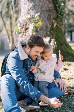 Elinde ladin dalı ve elinde koni olan küçük bir kız babasının kucağına oturup yere bakıyor. Yüksek kalite fotoğraf