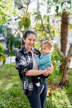 Küçük kız gülümseyen bir annenin kollarında oturur ve yeşil bir bahçedeki mesafeye bakar. Yüksek kalite fotoğraf