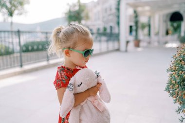 Küçük kız pembe bir oyuncak tavşana sarılıyor ve yeşil bir çalıya bakıyor. Yan görüş. Yüksek kalite fotoğraf