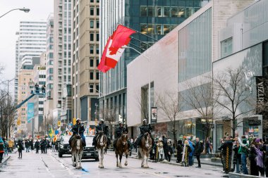 Bir geçit töreninde, atlı polis heyecanlı kalabalığın ve dekorasyonun ortasında Kanada bayrağını sergiliyor.