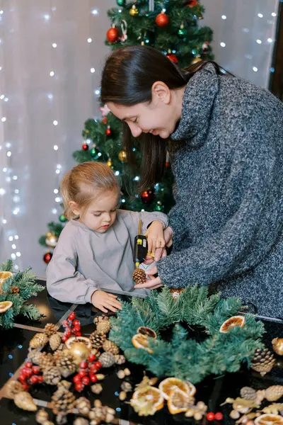 Küçük kız yeni yıl çelengi için annesinin elindeki kozalağa tabancadan tutkal sürüyor. Yüksek kalite fotoğraf
