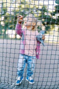 Küçük kız bir halat ağın arkasında duruyor, hücreyi eliyle tutuyor ve yukarı bakıyor. Yüksek kalite fotoğraf