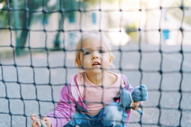 Tenis ağının arkasındaki kaldırımda çömelmiş oyuncağı olan küçük bir kız. Yüksek kalite fotoğraf