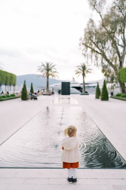Küçük kız uzun dikdörtgen bir göletin kenarında duruyor ve ağaçlara bakıyor. Arkadan bak. Yüksek kalite fotoğraf