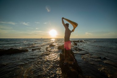 Genç bir adam gün batımında bir deniz sahilindeki bir rıhtımda gelişigüzel duruyor. Denizin üzerindeki ufuk.