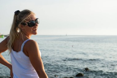 Genç, güzel, gülümseyen kadın gün batımında denizin kenarında duruyor.