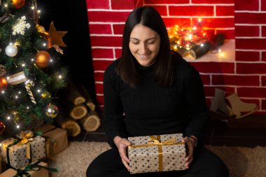 Yılbaşı hediyesini açan genç bir kadın Noel ağacı ve şöminenin önünde oturuyor. Paketler açılıyor 