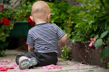 The boy is sitting on the ground in the yard and playing among the flowers