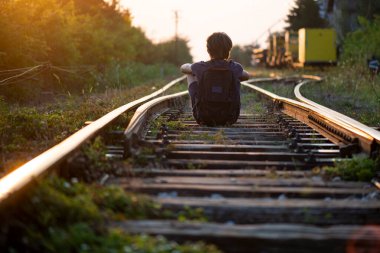 A boy with a backpack on his back sits on train tracks at dusk 