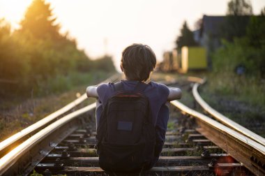 A boy with a backpack on his back sits on train tracks at dusk 