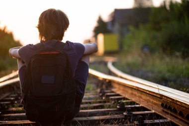 A boy with a backpack on his back sits on train tracks at dusk 