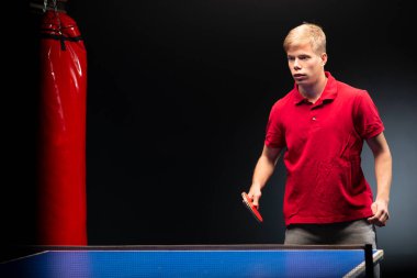 Portrait of a young man standing with a racket in his hand next to a table tennis table on a black background